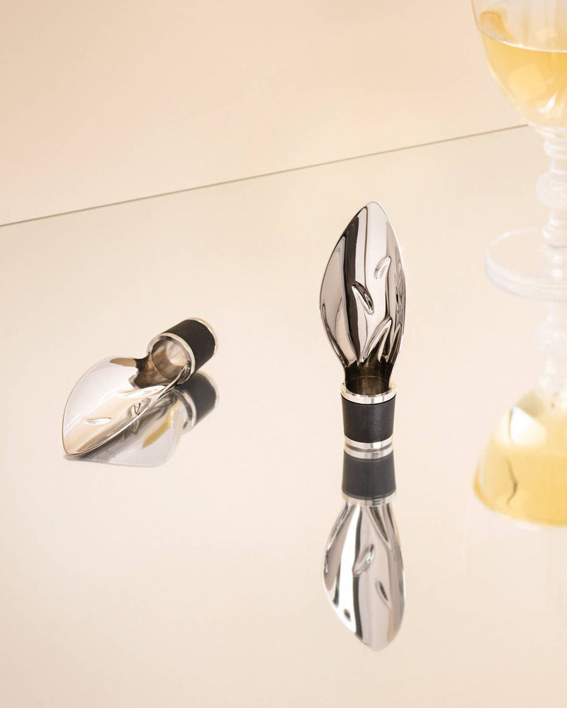 L'Atelier du Vin - Pourer-leaf stainless-steel aerators with rubber gaskets, shown on a mirrored surface beside a glass of white wine, enhancing aroma and pour control.