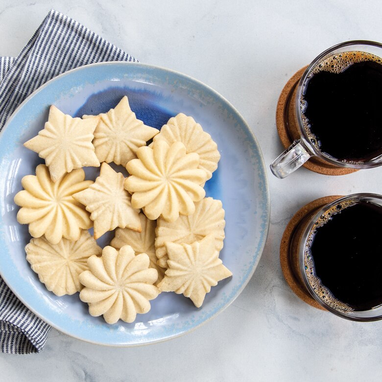 Nordic Ware | Pretty Pleated Cookie Stamps with star, diamond, and floral designs showcased on cookies, accompanied by two cups of coffee on a table.