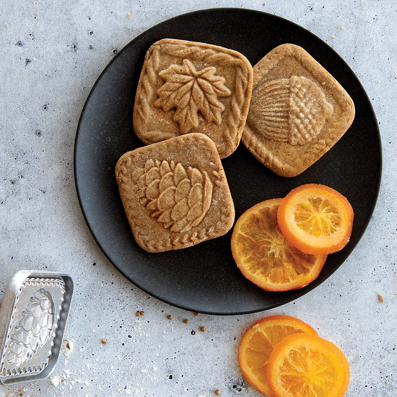 Nordic Ware Fall Forest Springerle Cookie Stamps on a plate with cookies and orange slices, showcasing autumn-inspired designs for elegant seasonal baking.