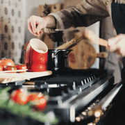Hand sprinkling salt from the Emile Henry | Burgundy Salt Pig into a simmering pot, with ripe tomatoes and kitchen utensils in the background.