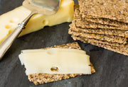 Norwegian Baked - Chili&Coriander Knekkebrød topped with Swiss cheese, accompanied by a cheese slicer and wedge, showcasing its fiber-rich, vegan, organic qualities.