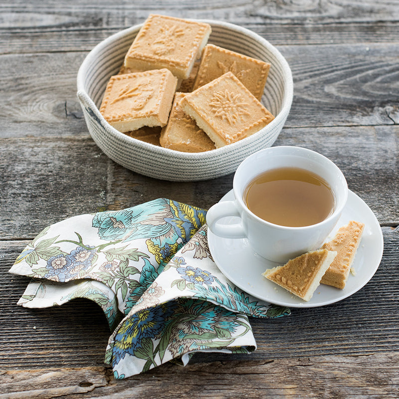 Nordic Ware English Shortbread Pan with baked shortbread cookies, showcasing intricate flower and fruit designs, accompanied by a cup of tea on a table.