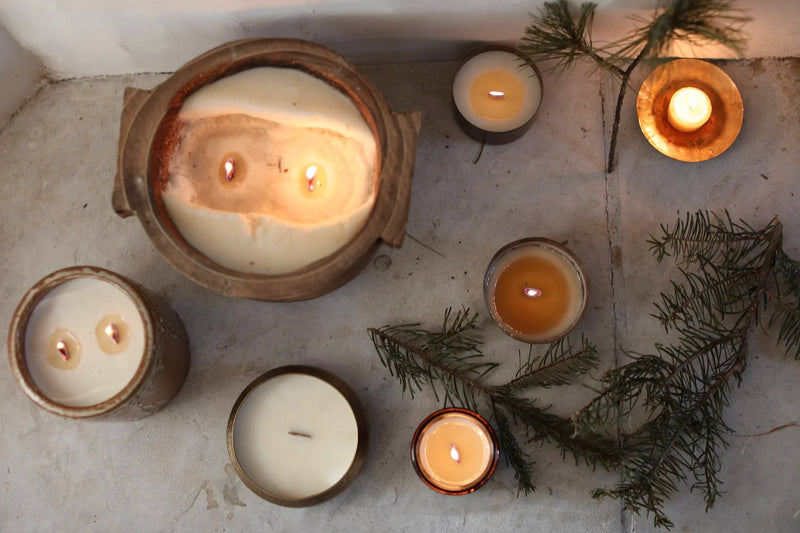 Among the Flowers | Essential Oil Candle, Through the Woods, rustic display with wooden wicks in jars, surrounded by evergreen sprigs on stone, enhancing a natural, festive ambiance.