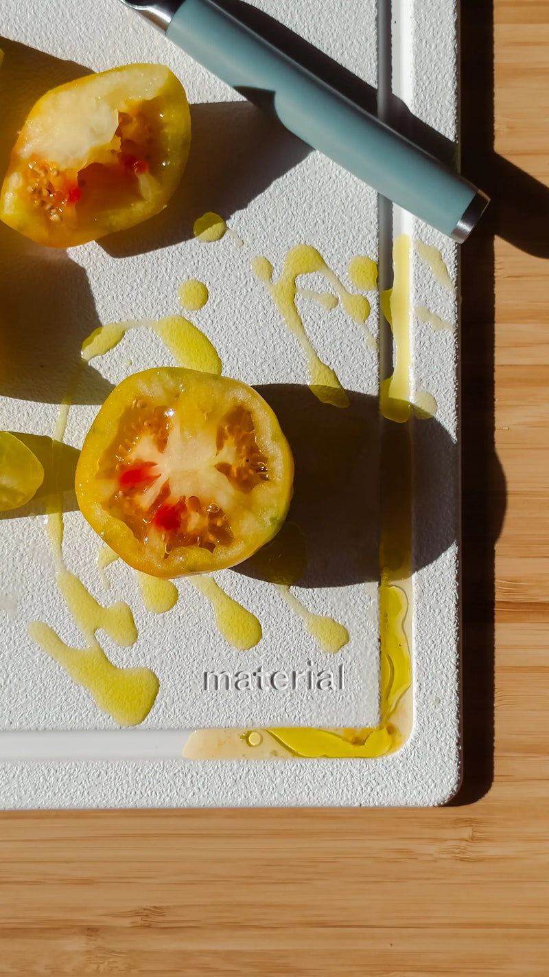Material - The MK Free Board: Ocean with a teal-handled utensil and halved yellow tomato on a wooden countertop, showcasing its non-porous, plant-based design for safe chopping.