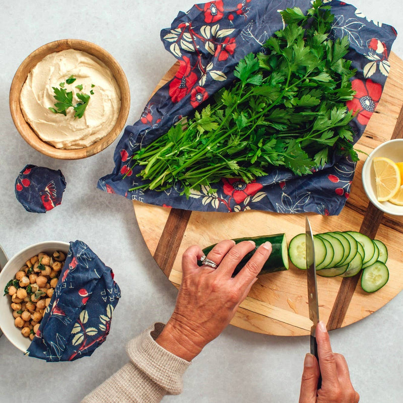 Hands slicing cucumber on a wooden board beside Bee's Wrap - Bread Wrap - Botanical Print, showcasing its use in preserving Mediterranean-style ingredients.