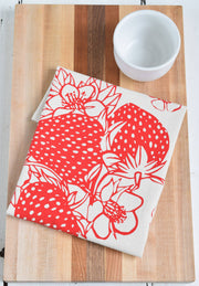 Hearth and Harrow - Strawberry Tea Towel, organic cotton, features a red fruit print design, displayed folded on a wooden surface with a white cup nearby.