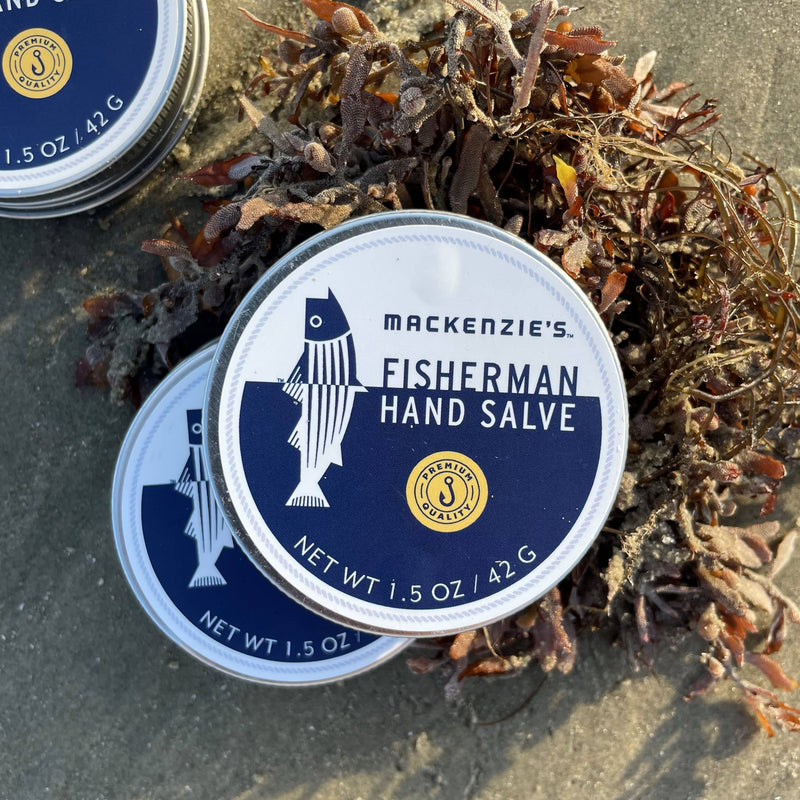 MacKenzie's Fisherman Hand Salve tins on sandy ground with dried kelp, offering serious dry skin protection with lemon scent. Ideal for fishermen, crafted in New England.