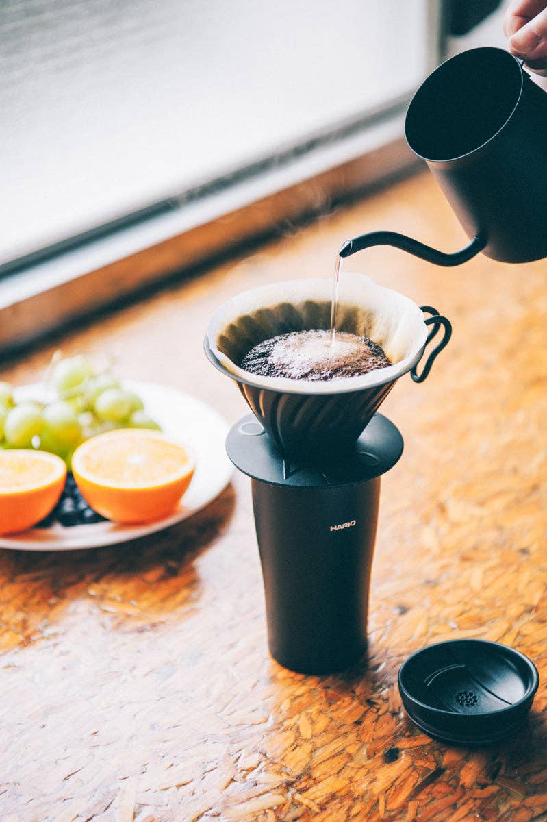 HARIO USA - V60 Metal Dripper: Silver, on a black travel mug, with a gooseneck kettle pouring over coffee grounds, highlights precision brewing and sleek design.