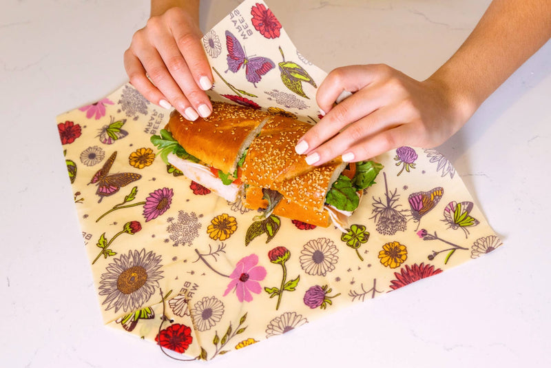 Bee's Wrap - Sandwich Wrap - Meadow Magic being folded around a deli sandwich on a countertop, showcasing its floral design and secure wooden bee button closure.