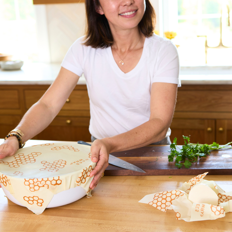 Woman in a kitchen holding a Bee's Wrap - Assorted 5 Pack, showcasing reusable wraps ideal for kitchen storage, made from sustainable materials.