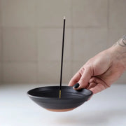 Hand holding a Gravesco Pottery - Incense Burner in black ceramic, showcasing its terracotta base and upright incense stick, ideal for catching ash elegantly.