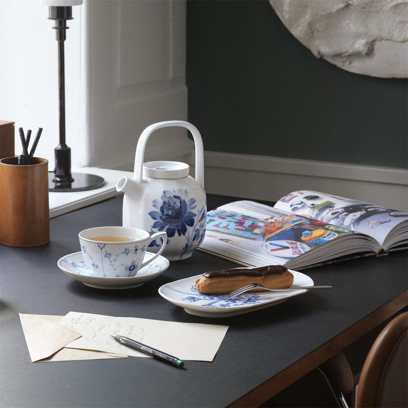 Royal Copenhagen | Blue Fluted Plain Cup & Saucer 28cl on a serene tea break setup with a teapot, éclair, book, and stationery on a tidy tabletop.