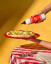Brightland - Pizza Oil being drizzled over a wood-fired pizza with zucchini ribbons and cheese, set on a red-and-white checkered tablecloth with a pizza cutter.