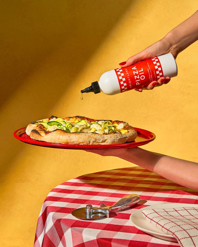 Brightland - Pizza Oil being drizzled over a wood-fired pizza with zucchini ribbons and cheese, set on a red-and-white checkered tablecloth with a pizza cutter.