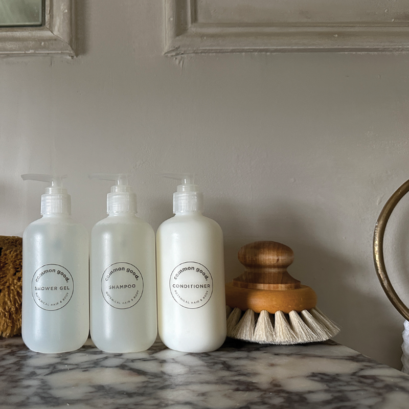 Three clear bottles with white pump tops labeled "Shower Gel," "Common Good | Botanical Shampoo & Conditioner," are on a marble countertop. A wooden brush with white bristles is next to the bottles. In the background, part of a circular metal handle and framed wall decor are visible, highlighting Common Good's appeal.