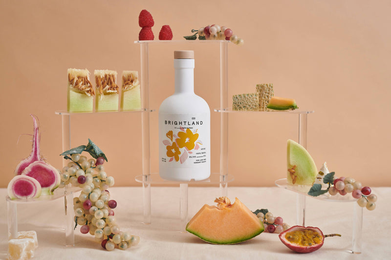 Brightland - ARISE Basil Infused Olive Oil bottle with a melon slice and passion fruit, showcasing its premium culinary appeal for Athens Cooks' customers.