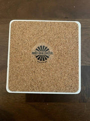 “Drinks on Me - COASTER Threw Out Back” features a cork surface with a sunburst logo, offering humor and charm for any bar or kitchen setting.