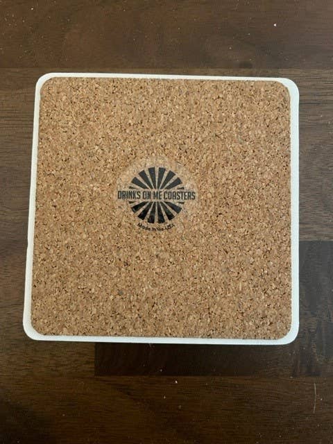 “Drinks on Me - COASTER Threw Out Back” features a cork surface with a sunburst logo, offering humor and charm for any bar or kitchen setting.