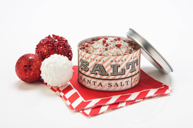 Beautiful Briny Sea - Santa Salt tin with coarse sea salt and pink peppercorns, atop striped napkins, next to three holiday baubles. Ideal for festive and versatile cooking.