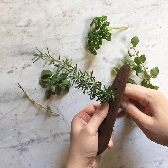 Hands using the Collin Garrity - Herb Stripper: Maple (Limited-release) to remove rosemary leaves, surrounded by parsley, oregano, and thyme on a marble countertop.