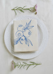 Hearth and Harrow - Wild Blueberry Tea Towel, organic cotton, featuring a blue-violet floral design, laid on a plate, showcasing its elegant, eco-friendly craftsmanship.