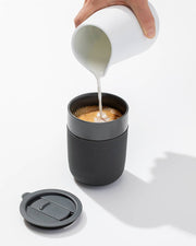 Person pouring milk into W&P - Reusable Ceramic Mug: Terrazzo Blush, featuring a splash-resistant lid and silicone sleeve, ideal for eco-friendly coffee enjoyment.