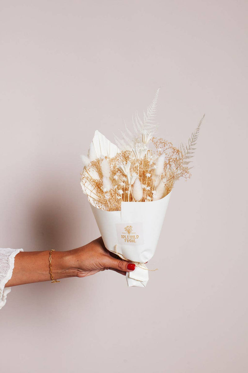 Idlewild Floral Co. - Snow Dried Flower Bouquet held by hand with red nails and a gold bracelet, wrapped in white paper, showcasing delicate preserved florals.