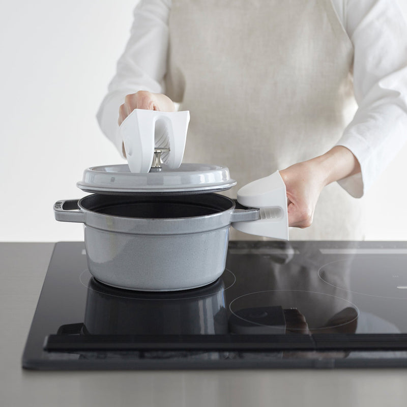 Person using Yamazaki Home Magnetic Pot Holders, Silicone, Black, to lift a cooking pot lid on a smooth-top stove, showcasing ease and safety in kitchen tasks.