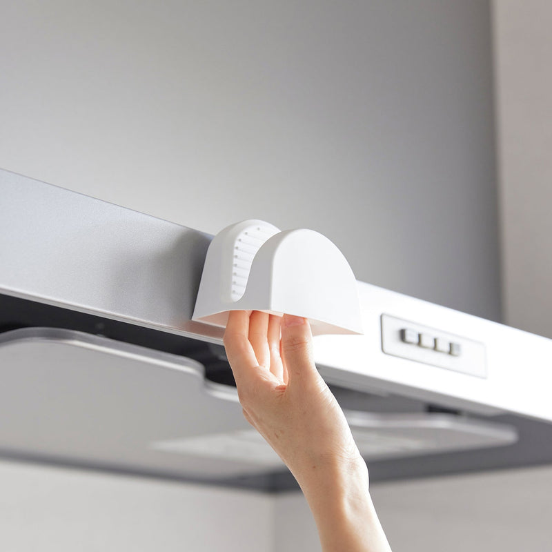 A hand attaches a Yamazaki Home Magnetic Pot Holder, Silicone, Black, to a range hood, ideal for hanging lightweight kitchen items.