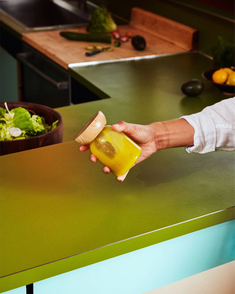 Hand holding Holcomb Studio Totter Salad Dressing Shaker, with a bowl of salad nearby, showcasing a mustard-like yellow liquid inside the jar.