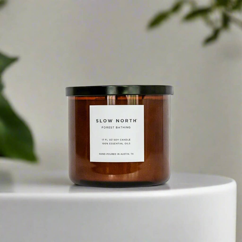 Slow North - Forest Bathing 3-Wick Non-Toxic Soy Candle with essential oils in a brown glass jar, black lid, surrounded by soft-focus greenery, invoking a serene, earthy ambiance.