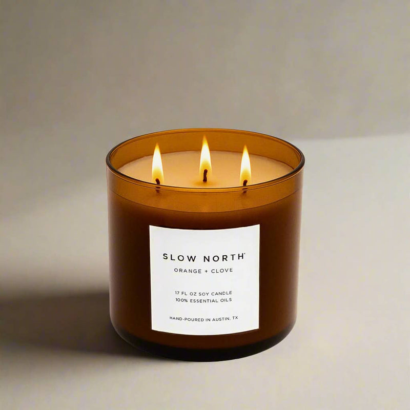 Slow North - Orange + Clove - Signature 3-Wick Non-Toxic Candle in amber glass jar, lit, emitting a cozy glow; hand-poured with essential oils, 17 oz.