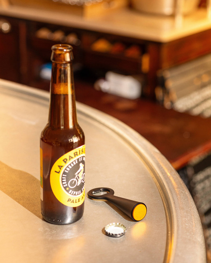 L'Atelier du Vin - USA - Bottle Opener - LE BRASSEUR – BLACK beside a La Parisienne Pale Ale bottle on a metallic tabletop.
