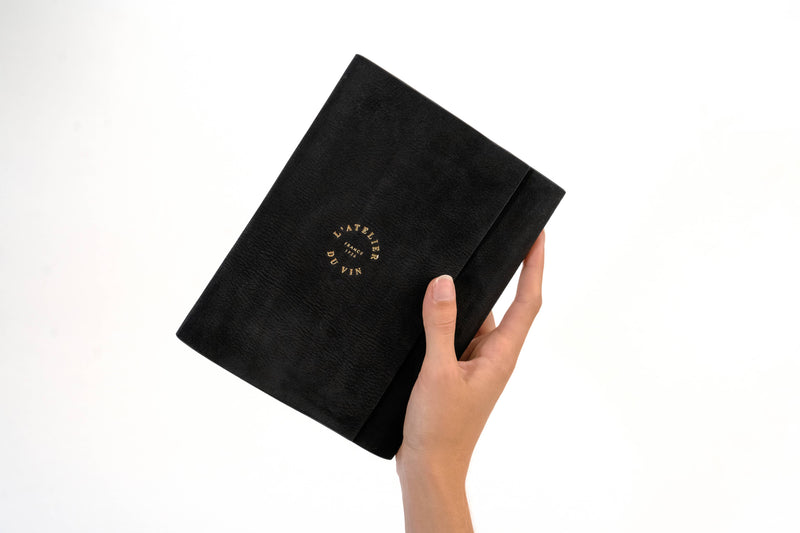 Hand holding a black leather L'ATELIER DU VIN notebook, embodying the Nomad Collection's elegance, perfect for gifting or special occasions.