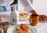 Brightland - ALIVE Extra Virgin Olive Oil bottle amidst a breakfast scene with yogurt, stone fruit, a wooden mug, and tomato-topped toast on a kitchen counter.