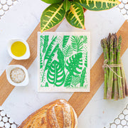Bespoke Provisions - Tropical Leaves Swedish Dishcloth on a green and white towel, surrounded by asparagus, bread, and bowls of oil and spices.