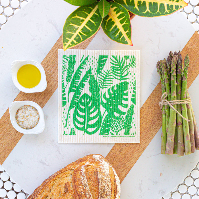 Bespoke Provisions - Tropical Leaves Swedish Dishcloth on a green and white towel, surrounded by asparagus, bread, and bowls of oil and spices.