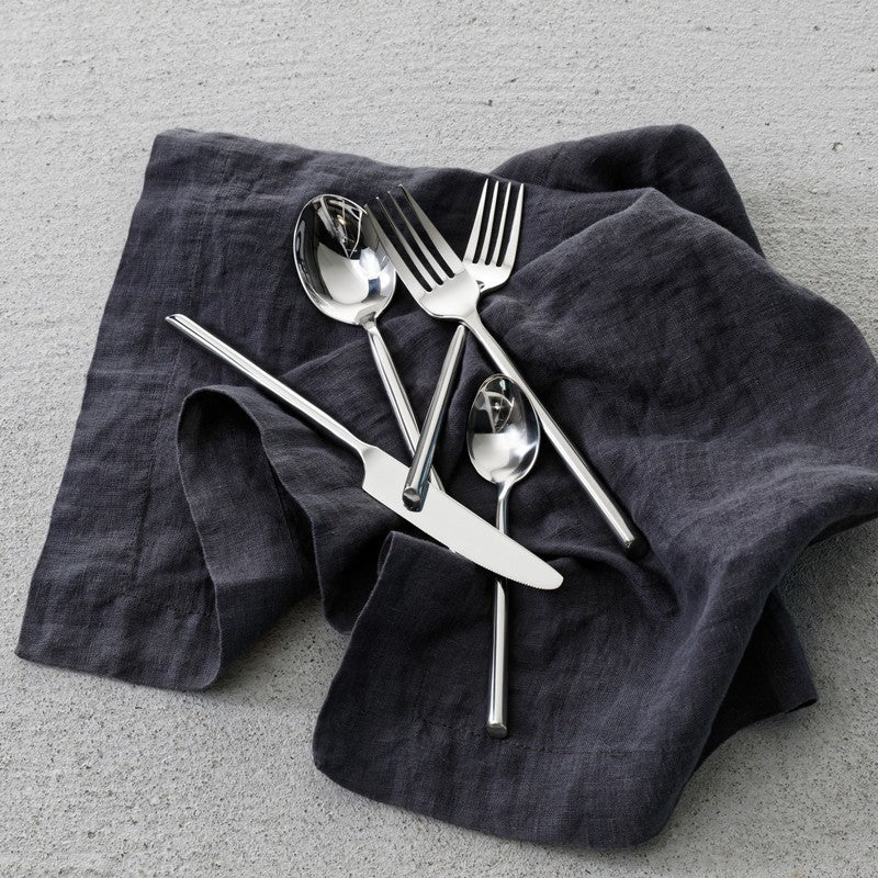 Zwiesel Fortessa Capri Flatware, 5-Piece Set, featuring polished stainless-steel knife, two forks, soup spoon, and teaspoon on a dark linen napkin.