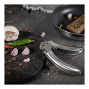 Kuhn Rikon | Epicurean Garlic Press on countertop beside cutting board with garlic, chilies, and herbs; ergonomic stainless steel design with easy-clean pullout sieve.