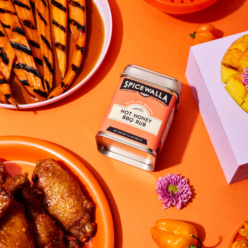 Spicewalla | Hot Honey BBQ Rub tin surrounded by grilled carrots, glazed chicken wings, habaneros, mango pieces, and a flower, highlighting its culinary versatility.