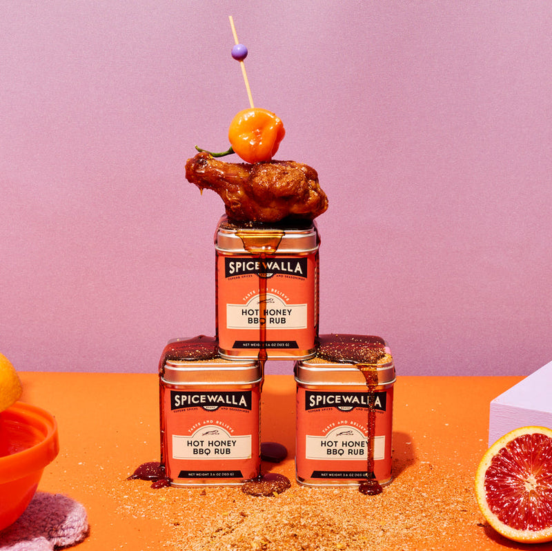 Spicewalla | Hot Honey BBQ Rub tins stacked with a glazed chicken wing and pepper skewer on top; sauce dripping, surrounded by rub, sauce bowl, and citrus half.