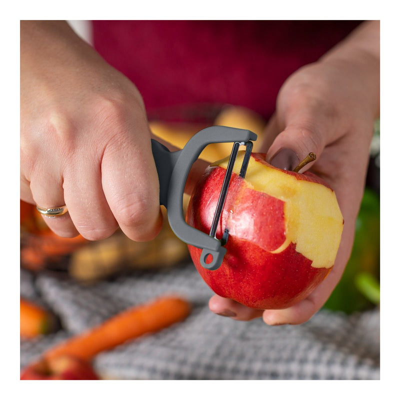 Close-up of hands using Kuhn Rikon | Original Swiss Peeler - Grey to peel a red apple, showcasing its razor-sharp carbon steel blades and ergonomic design.