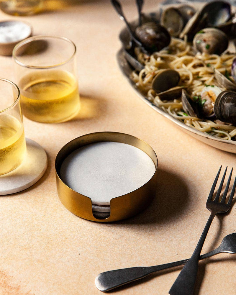 Farmhouse Pottery - Marble Laurel Coasters in a brass holder, surrounded by lowball glasses and a pasta dish, showcasing elegant surface protection.