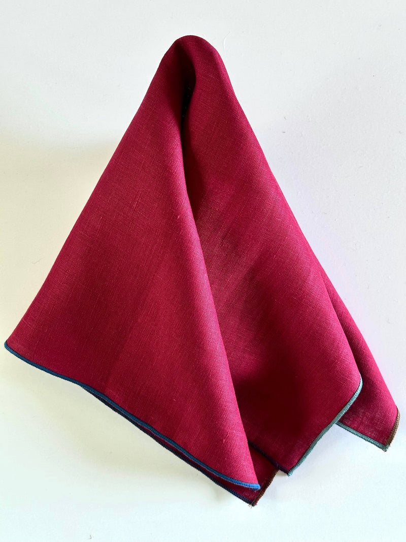Tisch New York - Napkins, Cranberry Linen (Night Fall Hem) featuring a red fabric with a multi-colored trim, draped in a pointed tent shape.