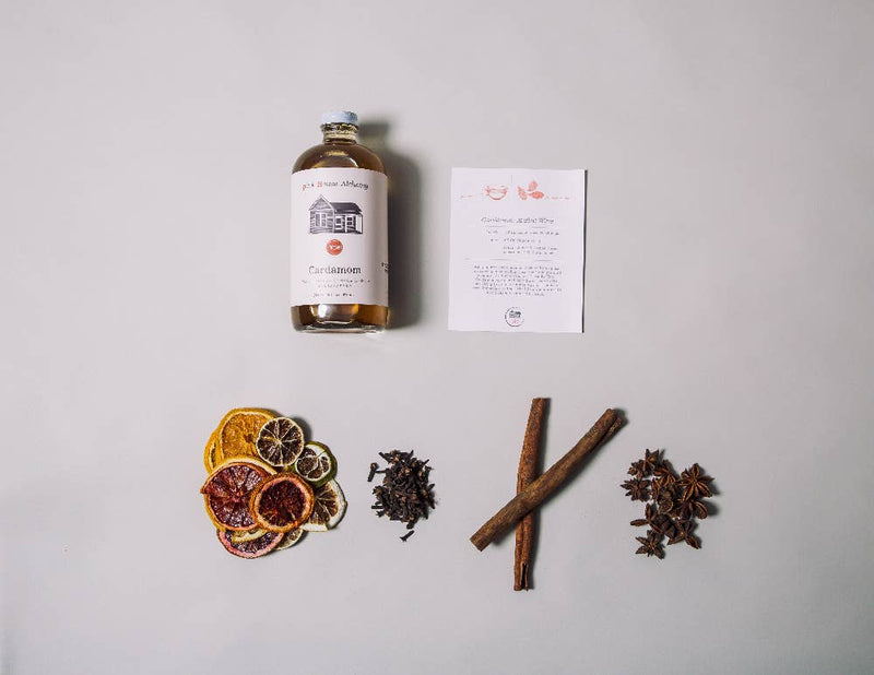 Pink House Alchemy - Cardamom Simple Syrup bottle surrounded by dried orange slices, cinnamon sticks, star anise, and cloves, emphasizing its culinary and aromatic use.