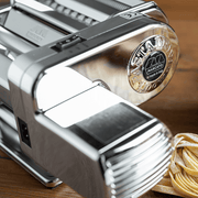 Marcato Atlas PastaDrive Motor 110V Premium on wooden surface, showcasing chrome-plated housing and dough thickness adjustment knob, with freshly cut, flour-dusted pasta beside it.