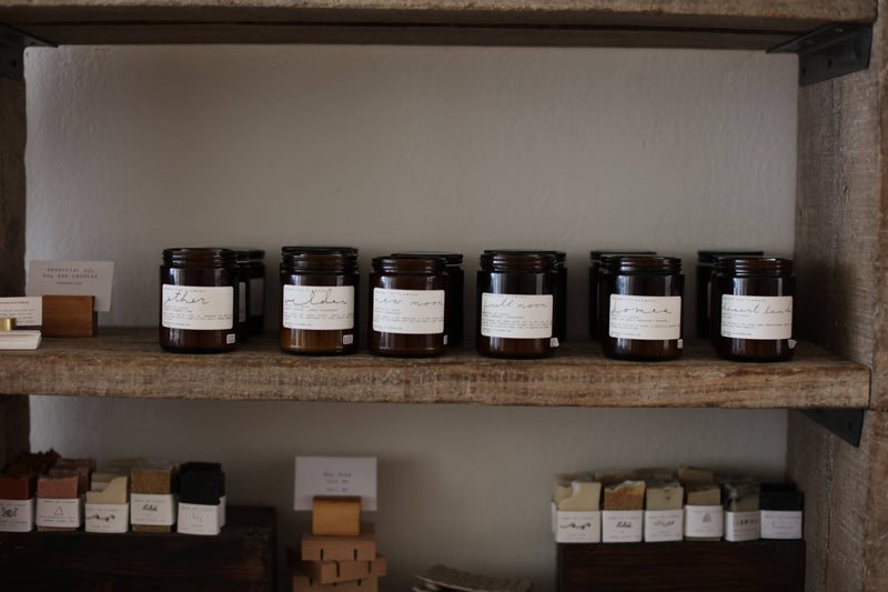 Among the Flowers - Essential Oil + Soy Wax Candle: Sitka, displayed on a shelf amidst various jars, featuring chamomile, bergamot, sage, and cedar scents.