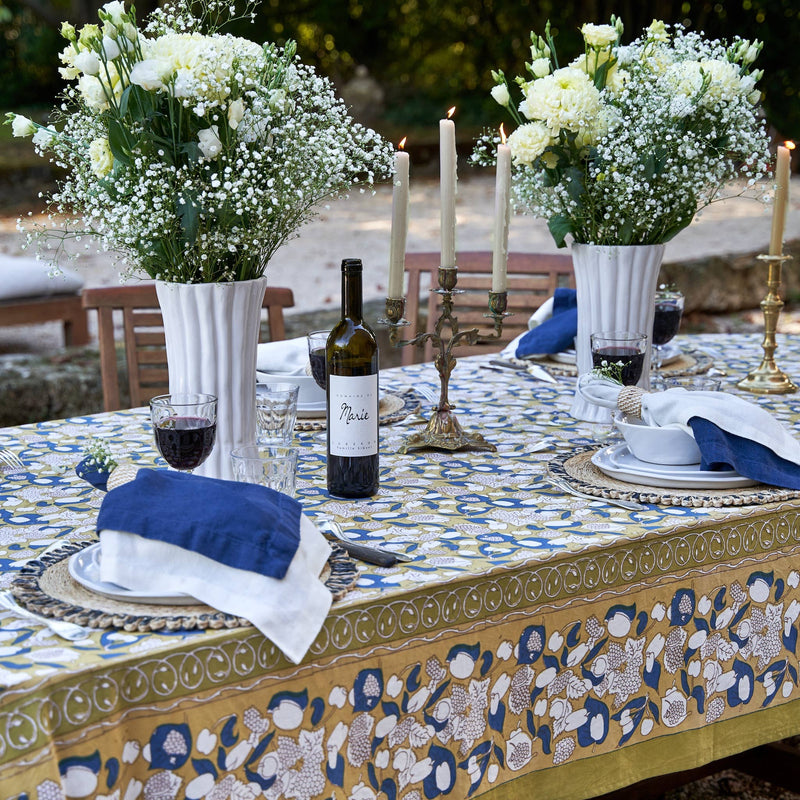 Couleur Nature - Forest Harvest Mustard & Blue tablecloth on an outdoor dining table, elegantly set with floral arrangements, candles, wine, and dinnerware.