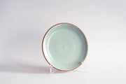 RachaelPots - Salad Plate: Nude/White, a handmade ceramic plate with a speckled light turquoise glaze and unglazed rim, displayed on a clear stand.