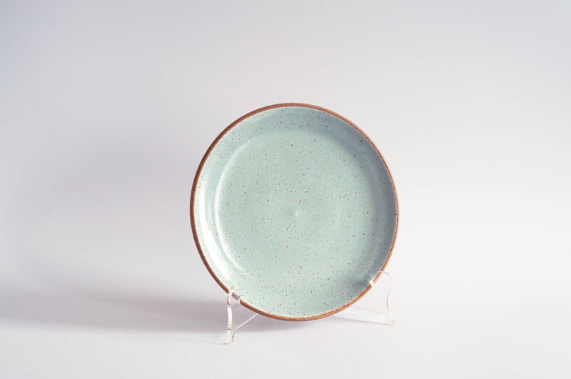 RachaelPots - Salad Plate: Nude/White, a handmade ceramic plate with a speckled light turquoise glaze and unglazed rim, displayed on a clear stand.